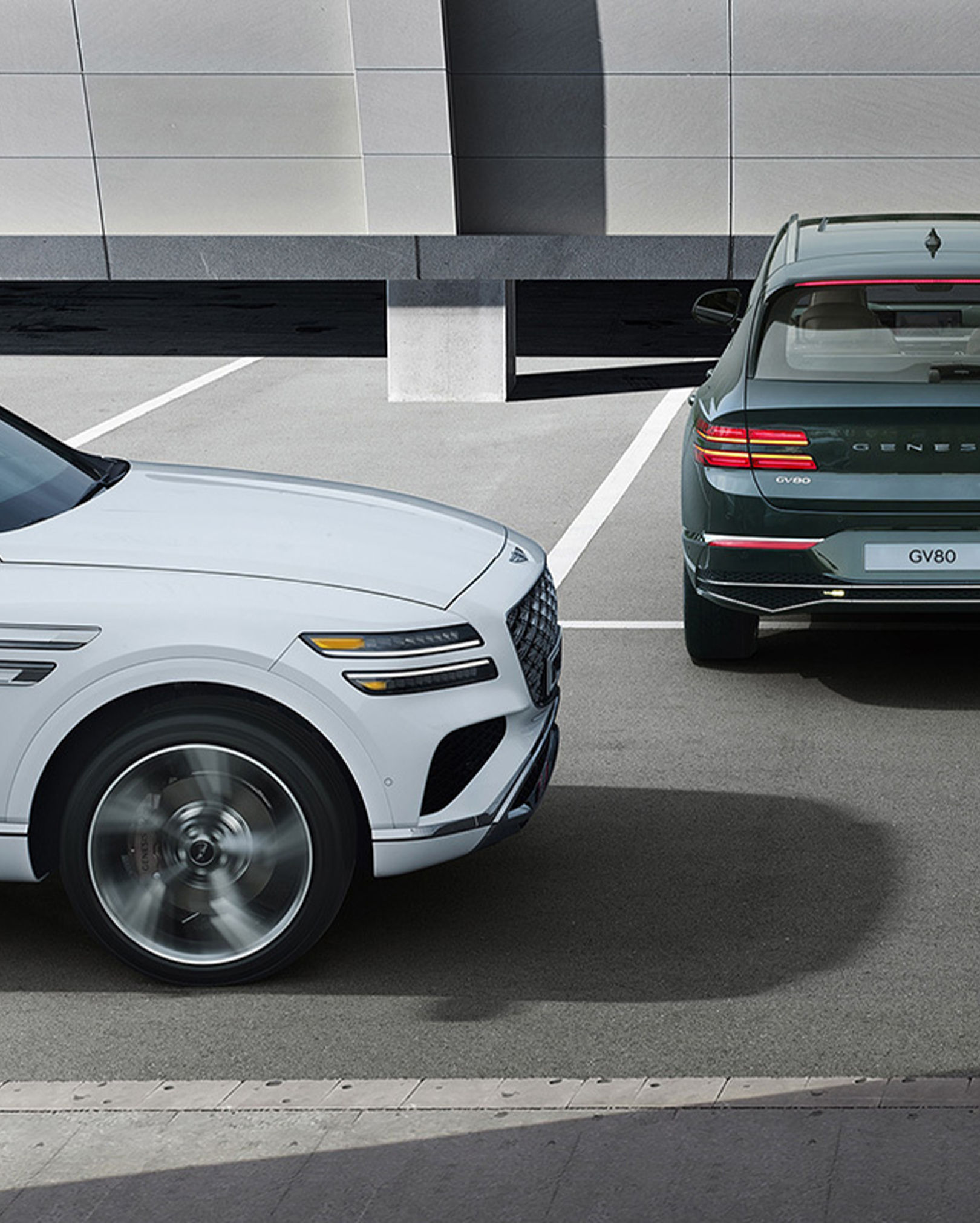 Storr Green GV80 Coupe is backing out of a parking spot, and a Uyuni White GV80 Coupe passes behind the vehicle on the left.