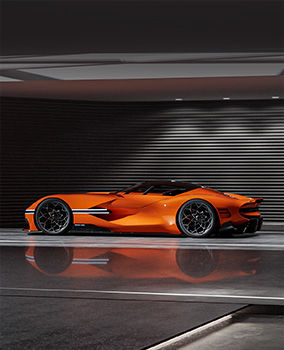 An orange-hued Genesis X Gran Berlineta Concept vehicle is displayed indoors, illuminated by white lights that highlight its low and sleek sports car body. The metallic interior flooring distinctly reflects the vehicle's body.