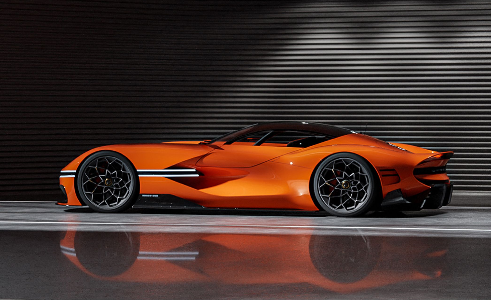 The X Gran Berlinetta Concept stands poised in front of a dark panel wall, illuminated by white overhead lights. Its side profile and lit headlamps reflect elegantly off the sleek, glossy floor.