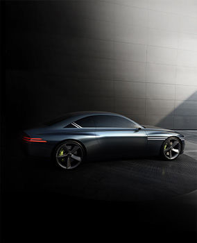 A side view of the Genesis X Concept vehicle parked in a darkened interior, illuminated by natural light streaming in from the upper right of the room. This light reflects off the bodywork, emphasizing the metallic color of the car's surface.