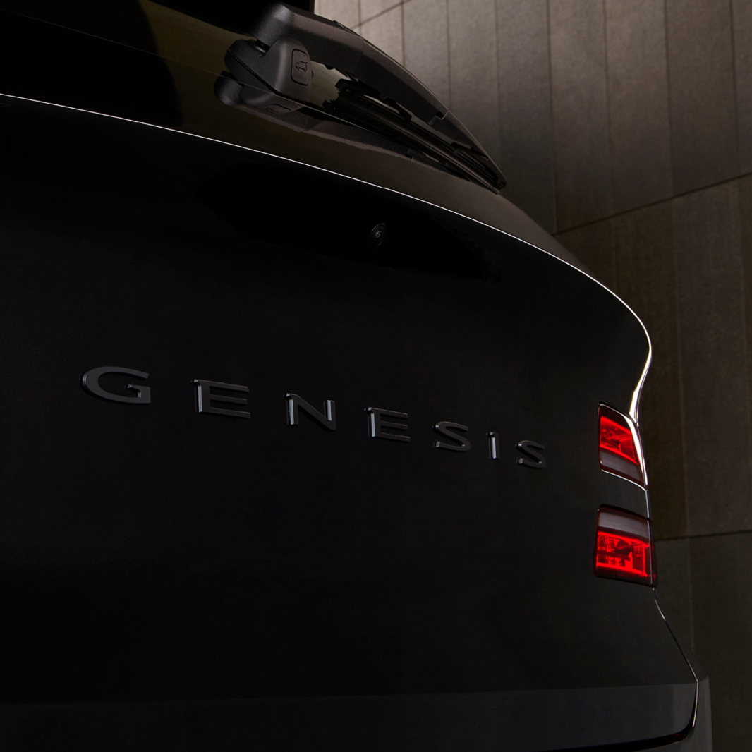 At the rear of the GV80 Black, the metallic lettering that spells 'GENESIS' is visible. Two red light strips are illuminated.
