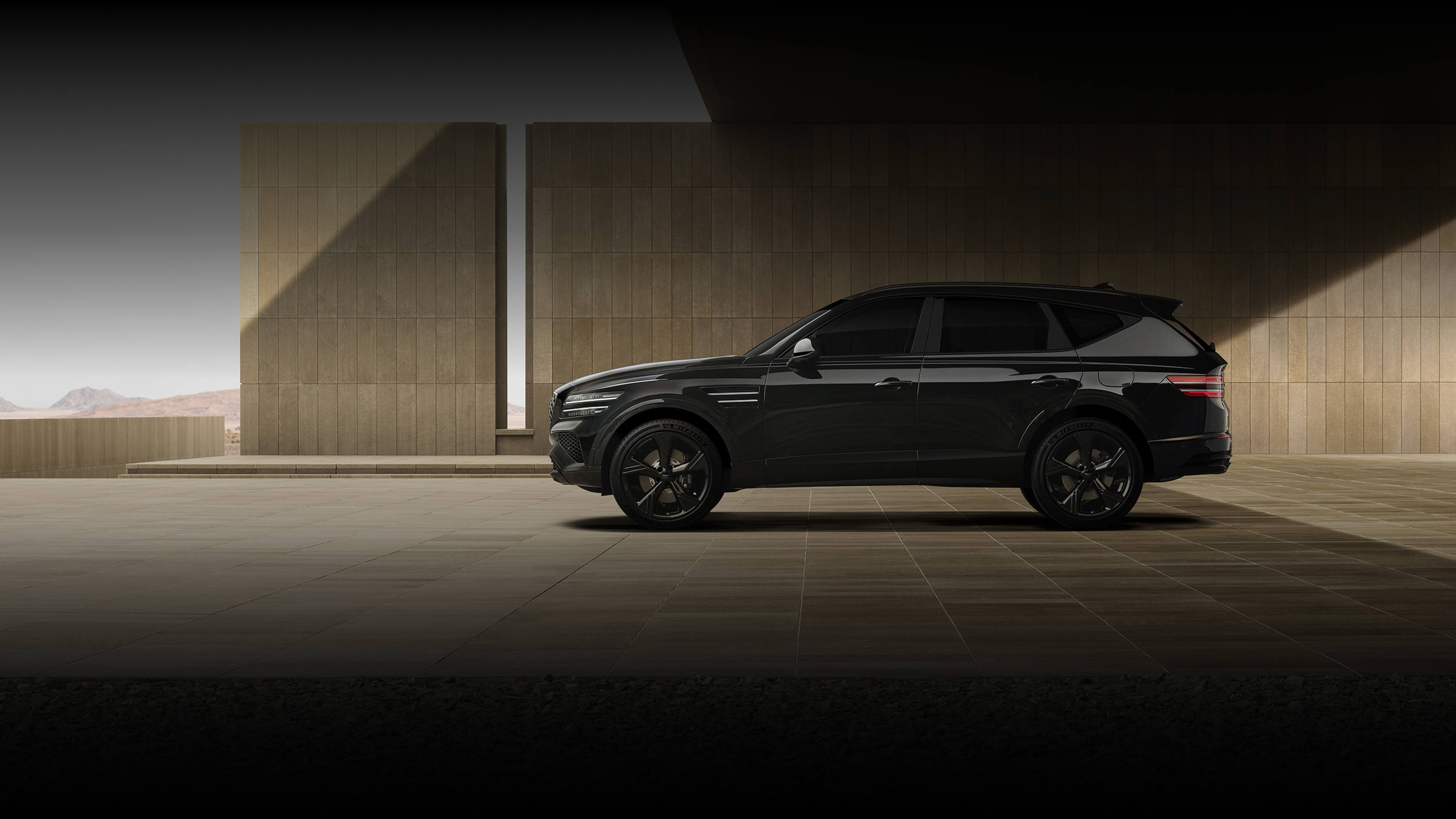 The GV80 Black is parked with its left side visible, in front of a high, grayish wall. The shadow cast by the high ceiling above makes the scene appear darker overall.