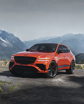 An orange-hued Genesis GV80 Coupe Concept vehicle is parked on a road with rugged mountains in the background. The headlights are on, and the front wheels are slightly turned with skid marks visible.