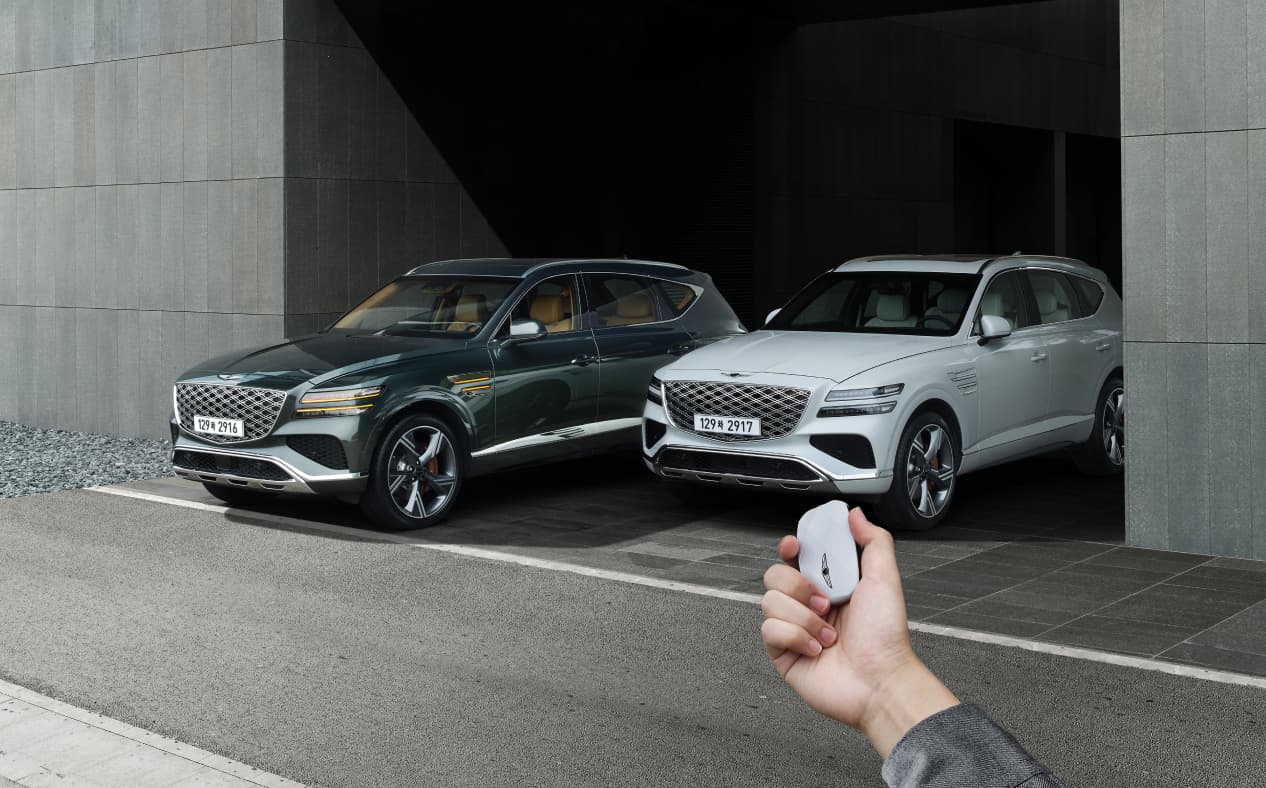 A Vik Black GV80 and a Uyuni White GV80 are parked, and the driver is operating the smart key outside the vehicle.