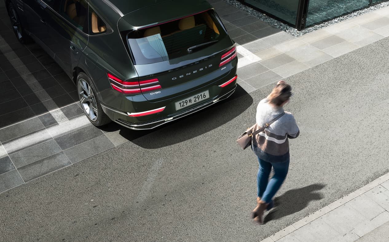 Storr Green GV80 backing out of a parking lot, and a woman is passing behind the vehicle.