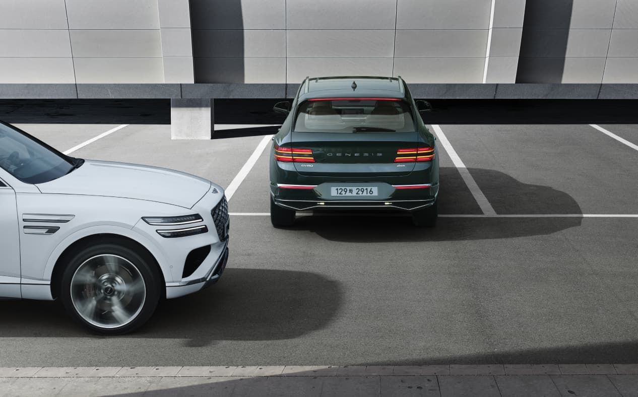 Storr Green GV80 is backing out of a parking spot, and a Uyuni White GV80 passes behind the vehicle on the left.